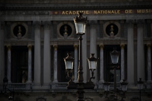 Several ornate street lamps stand in front of a classical building with tall columns and decorative sculptures. The architecture indicates a historic style with a focus on grandeur and symmetry. The building has the phrase 'Academie Nationale de Musique' inscribed at the top.