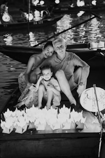 A family of three sits together on a boat at night, with a bright lantern illuminating their faces. Decorative paper lanterns float on the water surface surrounding them, and other boats can be seen in the background, creating a serene and festive atmosphere.