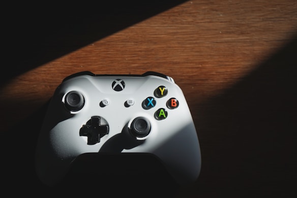 A white gaming controller with black buttons and colored symbols sits on a wooden surface. Sunlight casts a partial shadow across the controller, highlighting the buttons labeled with the letters X, Y, B, and A in blue, yellow, red, and green respectively.