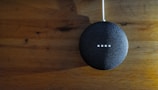 A small, round, fabric-covered smart speaker is positioned against a wooden surface. Four small white lights are illuminated on its surface, indicating activity or response.