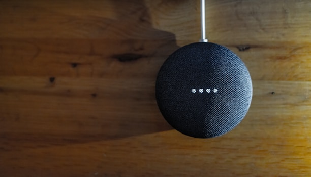 A small, round, fabric-covered smart speaker is positioned against a wooden surface. Four small white lights are illuminated on its surface, indicating activity or response.