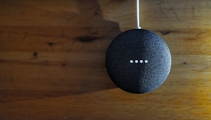 A small, round, fabric-covered smart speaker is positioned against a wooden surface. Four small white lights are illuminated on its surface, indicating activity or response.
