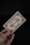 A hand holding a vintage-looking ticket with ornate borders and text indicating 'London to Hogwarts' and 'Platform 9¾'. The ticket has a sepia tone, giving it an antique feel, against a dark black background.