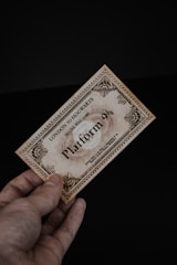 A hand holding a vintage-looking ticket with ornate borders and text indicating 'London to Hogwarts' and 'Platform 9¾'. The ticket has a sepia tone, giving it an antique feel, against a dark black background.