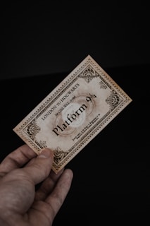 A hand holding a vintage-looking ticket with ornate borders and text indicating 'London to Hogwarts' and 'Platform 9¾'. The ticket has a sepia tone, giving it an antique feel, against a dark black background.
