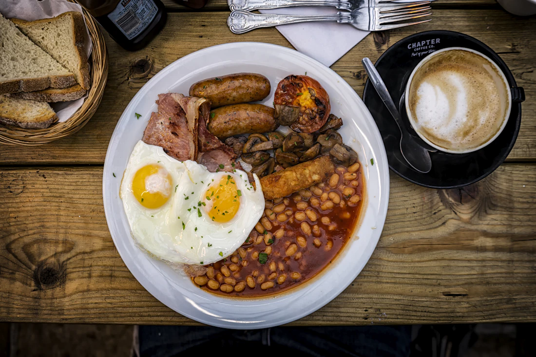 A generous full English breakfast on a rustic table: eggs, sausages, beans, grilled tomato and toast beside a cup of coffee