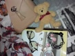 A neatly arranged collection of fashion accessories, including a beige handbag with a metallic clasp and tassel, a small plush teddy bear with a burgundy ribbon, and a pair of black-framed eyeglasses. The items rest on a textured background with floral patterns and typography. Several pieces of jewelry, such as rings, earrings, and a black bracelet, are placed on a magazine with a glossy page featuring more accessories.