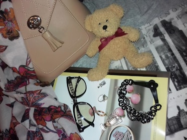 A neatly arranged collection of fashion accessories, including a beige handbag with a metallic clasp and tassel, a small plush teddy bear with a burgundy ribbon, and a pair of black-framed eyeglasses. The items rest on a textured background with floral patterns and typography. Several pieces of jewelry, such as rings, earrings, and a black bracelet, are placed on a magazine with a glossy page featuring more accessories.