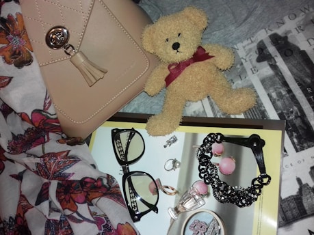 A neatly arranged collection of fashion accessories, including a beige handbag with a metallic clasp and tassel, a small plush teddy bear with a burgundy ribbon, and a pair of black-framed eyeglasses. The items rest on a textured background with floral patterns and typography. Several pieces of jewelry, such as rings, earrings, and a black bracelet, are placed on a magazine with a glossy page featuring more accessories.