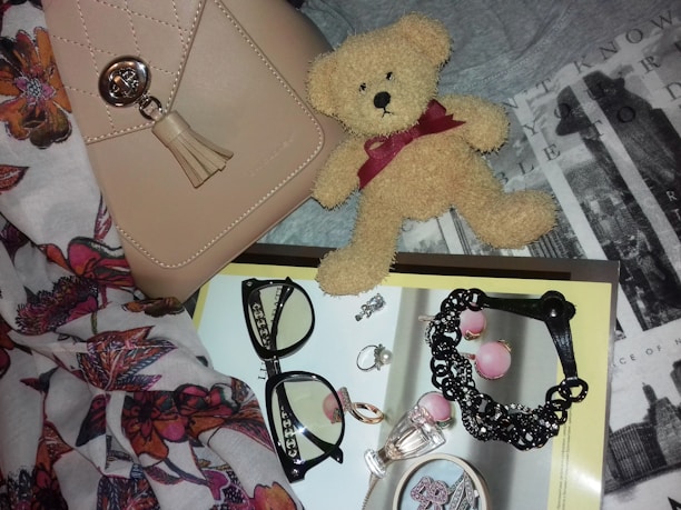 A neatly arranged collection of fashion accessories, including a beige handbag with a metallic clasp and tassel, a small plush teddy bear with a burgundy ribbon, and a pair of black-framed eyeglasses. The items rest on a textured background with floral patterns and typography. Several pieces of jewelry, such as rings, earrings, and a black bracelet, are placed on a magazine with a glossy page featuring more accessories.
