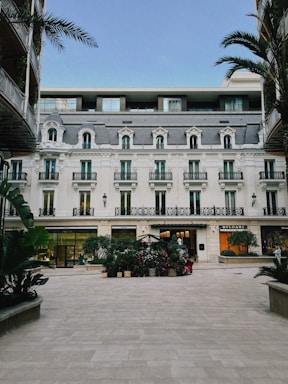 A beautiful building with classic European architecture featuring symmetrical windows and ornate balconies. The foreground includes a courtyard with potted plants and two high-end store signs on the ground floor, Bvlgari and another luxury brand. Palm trees frame the building on both sides, and the sky is clear blue.
