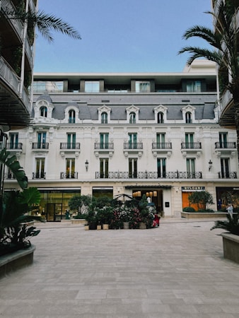 A beautiful building with classic European architecture featuring symmetrical windows and ornate balconies. The foreground includes a courtyard with potted plants and two high-end store signs on the ground floor, Bvlgari and another luxury brand. Palm trees frame the building on both sides, and the sky is clear blue.