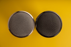 Two circular speaker grilles with a metallic mesh pattern are placed side by side on a vibrant yellow background. The symmetrical arrangement creates a balanced composition, with the contrast of the dark grilles against the bright background drawing attention.