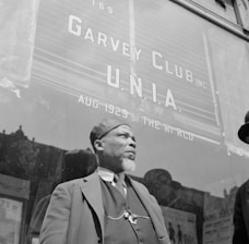 A man with a beard is standing in front of a building with large signage that reads 'Garvey Club U.N.I.A. Aug. 1929 of the World.' He is wearing a hat and a three-piece suit, with a chain visible on his vest. The background suggests an urban setting with a window reflecting the outside scene.