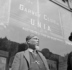 A man with a beard is standing in front of a building with large signage that reads 'Garvey Club U.N.I.A. Aug. 1929 of the World.' He is wearing a hat and a three-piece suit, with a chain visible on his vest. The background suggests an urban setting with a window reflecting the outside scene.