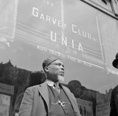 A man with a beard is standing in front of a building with large signage that reads 'Garvey Club U.N.I.A. Aug. 1929 of the World.' He is wearing a hat and a three-piece suit, with a chain visible on his vest. The background suggests an urban setting with a window reflecting the outside scene.