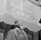 A man with a beard is standing in front of a building with large signage that reads 'Garvey Club U.N.I.A. Aug. 1929 of the World.' He is wearing a hat and a three-piece suit, with a chain visible on his vest. The background suggests an urban setting with a window reflecting the outside scene.