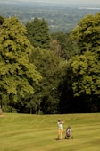 A golfer mid-swing on a lush, green golf course with a golf bag on a trolley nearby. The background features dense green trees and an expansive view of a distant valley under a clear sky.