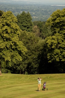 A golfer mid-swing on a lush, green golf course with a golf bag on a trolley nearby. The background features dense green trees and an expansive view of a distant valley under a clear sky.