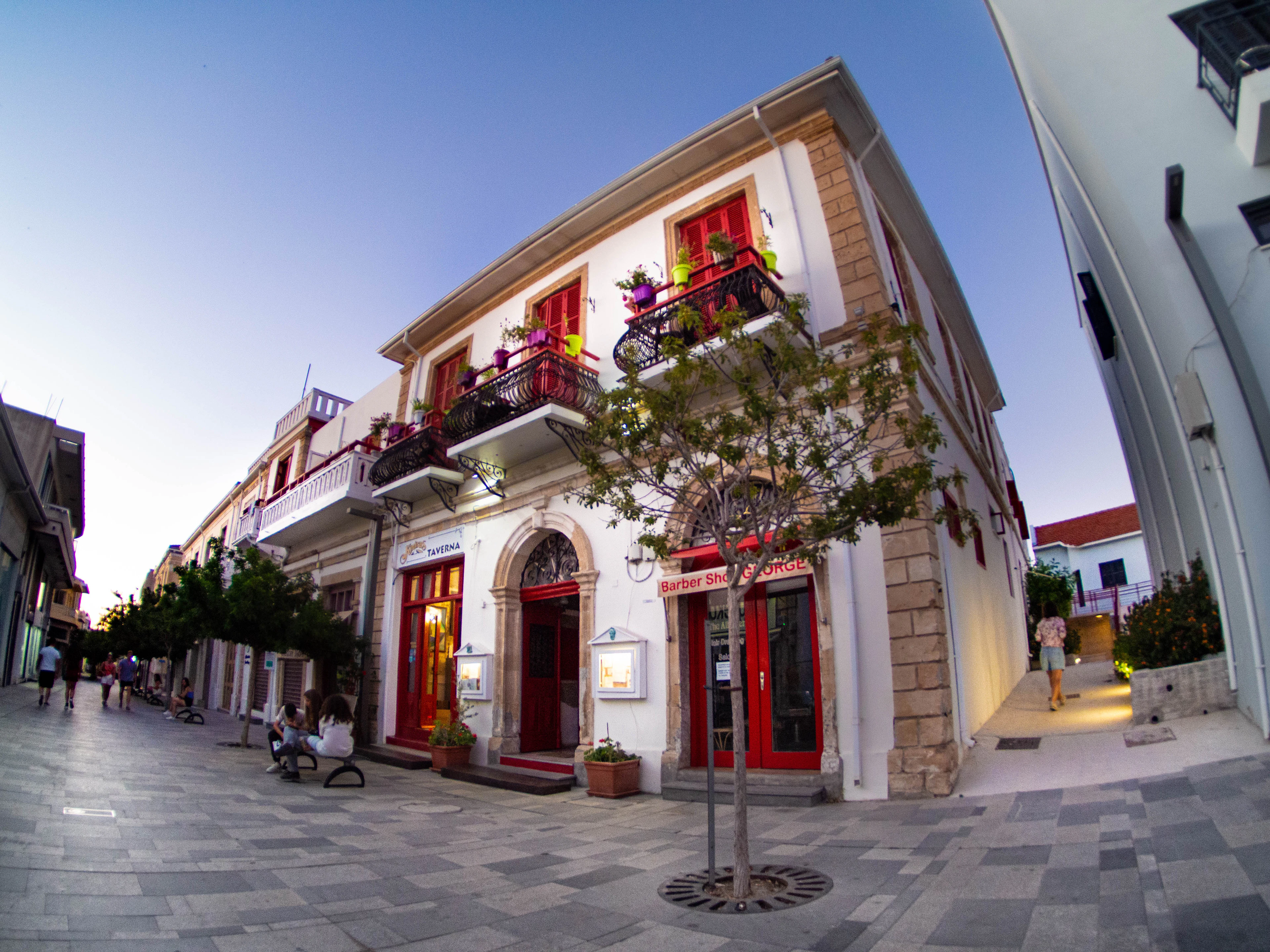 A pedestrianised street in the Old Town of Paphos at dusk, with a colonial-style building in white and red — shuttered windows, flower-filled balconies and lit shopfronts