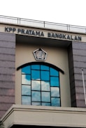 A building facade featuring a large arched window with multiple panes reflecting the blue sky and clouds. Above the window, there is a sign with the text 'KPP PRATAMA BANGKALAN' and an emblem in the center. The structure has gray stone-like columns on the sides and a cream-colored background.