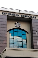 A building facade featuring a large arched window with multiple panes reflecting the blue sky and clouds. Above the window, there is a sign with the text 'KPP PRATAMA BANGKALAN' and an emblem in the center. The structure has gray stone-like columns on the sides and a cream-colored background.