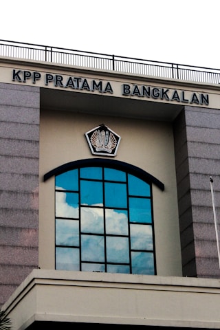 A building facade featuring a large arched window with multiple panes reflecting the blue sky and clouds. Above the window, there is a sign with the text 'KPP PRATAMA BANGKALAN' and an emblem in the center. The structure has gray stone-like columns on the sides and a cream-colored background.