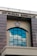 A building facade featuring a large arched window with multiple panes reflecting the blue sky and clouds. Above the window, there is a sign with the text 'KPP PRATAMA BANGKALAN' and an emblem in the center. The structure has gray stone-like columns on the sides and a cream-colored background.