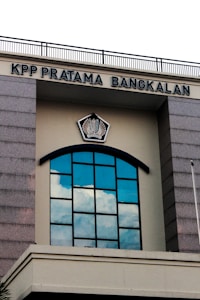 A building facade featuring a large arched window with multiple panes reflecting the blue sky and clouds. Above the window, there is a sign with the text 'KPP PRATAMA BANGKALAN' and an emblem in the center. The structure has gray stone-like columns on the sides and a cream-colored background.