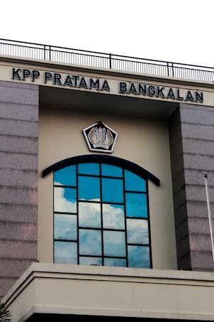 A building facade featuring a large arched window with multiple panes reflecting the blue sky and clouds. Above the window, there is a sign with the text 'KPP PRATAMA BANGKALAN' and an emblem in the center. The structure has gray stone-like columns on the sides and a cream-colored background.