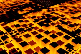 Abstract scene featuring a glowing geometric pattern of illuminated cubes and squares. The pattern is predominantly in shades of orange and black, creating a visually striking contrast. It resembles a digital or futuristic landscape, possibly representing a circuit board or a city at night.