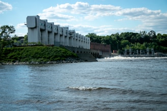 A large dam-like structure with multiple arches is situated beside a river. The building is modern and industrial in design, with a backdrop of lush, green trees under a partly cloudy sky.