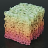 A complex, three-dimensional cube-shaped structure composed of interconnected, wireframe-like rectangular shapes. The structure features a gradient color scheme transitioning from shades of green at the top to yellow and pink at the bottom, set against a dark background.