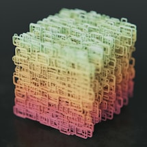 A complex, three-dimensional cube-shaped structure composed of interconnected, wireframe-like rectangular shapes. The structure features a gradient color scheme transitioning from shades of green at the top to yellow and pink at the bottom, set against a dark background.