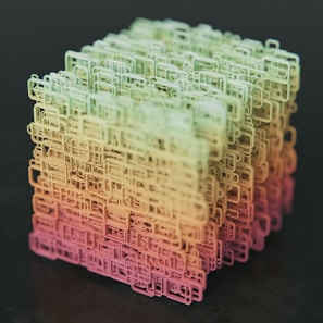 A complex, three-dimensional cube-shaped structure composed of interconnected, wireframe-like rectangular shapes. The structure features a gradient color scheme transitioning from shades of green at the top to yellow and pink at the bottom, set against a dark background.
