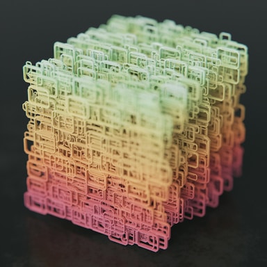 A complex, three-dimensional cube-shaped structure composed of interconnected, wireframe-like rectangular shapes. The structure features a gradient color scheme transitioning from shades of green at the top to yellow and pink at the bottom, set against a dark background.