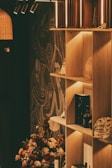 A warmly lit shelf with various decorative items including bottles and a bag, set against a wall with artistic patterns. A floral arrangement with earthy tones occupies the foreground, adding a natural touch.