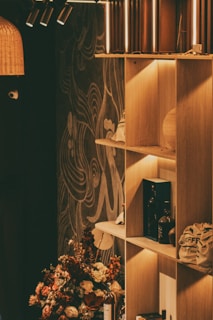 A warmly lit shelf with various decorative items including bottles and a bag, set against a wall with artistic patterns. A floral arrangement with earthy tones occupies the foreground, adding a natural touch.