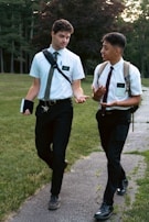 Two young men wearing white shirts, ties, and black pants are walking along a pathway in a park. They both have backpacks and are engaged in a conversation, gesturing as they talk. One is holding a small book in his hand. The background features lush green grass and tall trees.