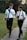 Two young men wearing white shirts, ties, and black pants are walking along a pathway in a park. They both have backpacks and are engaged in a conversation, gesturing as they talk. One is holding a small book in his hand. The background features lush green grass and tall trees.