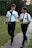 Two young men wearing white shirts, ties, and black pants are walking along a pathway in a park. They both have backpacks and are engaged in a conversation, gesturing as they talk. One is holding a small book in his hand. The background features lush green grass and tall trees.