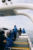 Passengers in blue jackets are standing and observing the scenery from a boat. The ocean surrounds the vessel, with rocky formations visible in the distance. The sky is overcast, contributing to a chilly atmosphere. A set of horns or speakers are mounted on the boat frame above.
