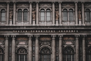 An ornate, neoclassical building facade featuring multiple columns, large windows, and decorative statues depicting historical figures. The inscription 'MAGYAR TUDOMÁNYOS AKADÉMIA' is prominently displayed above the entrance.
