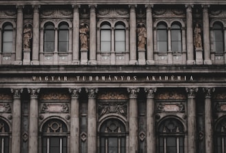 An ornate, neoclassical building facade featuring multiple columns, large windows, and decorative statues depicting historical figures. The inscription 'MAGYAR TUDOMÁNYOS AKADÉMIA' is prominently displayed above the entrance.