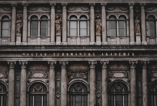 An ornate, neoclassical building facade featuring multiple columns, large windows, and decorative statues depicting historical figures. The inscription 'MAGYAR TUDOMÁNYOS AKADÉMIA' is prominently displayed above the entrance.
