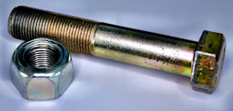 A metallic bolt and nut are placed on a white surface. The bolt has a hexagonal head and is positioned parallel to the nut, which is similarly hexagonal and threaded. Both objects have a shiny metallic surface with slight colorful reflections due to the lighting.