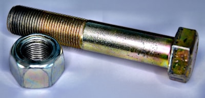 A metallic bolt and nut are placed on a white surface. The bolt has a hexagonal head and is positioned parallel to the nut, which is similarly hexagonal and threaded. Both objects have a shiny metallic surface with slight colorful reflections due to the lighting.