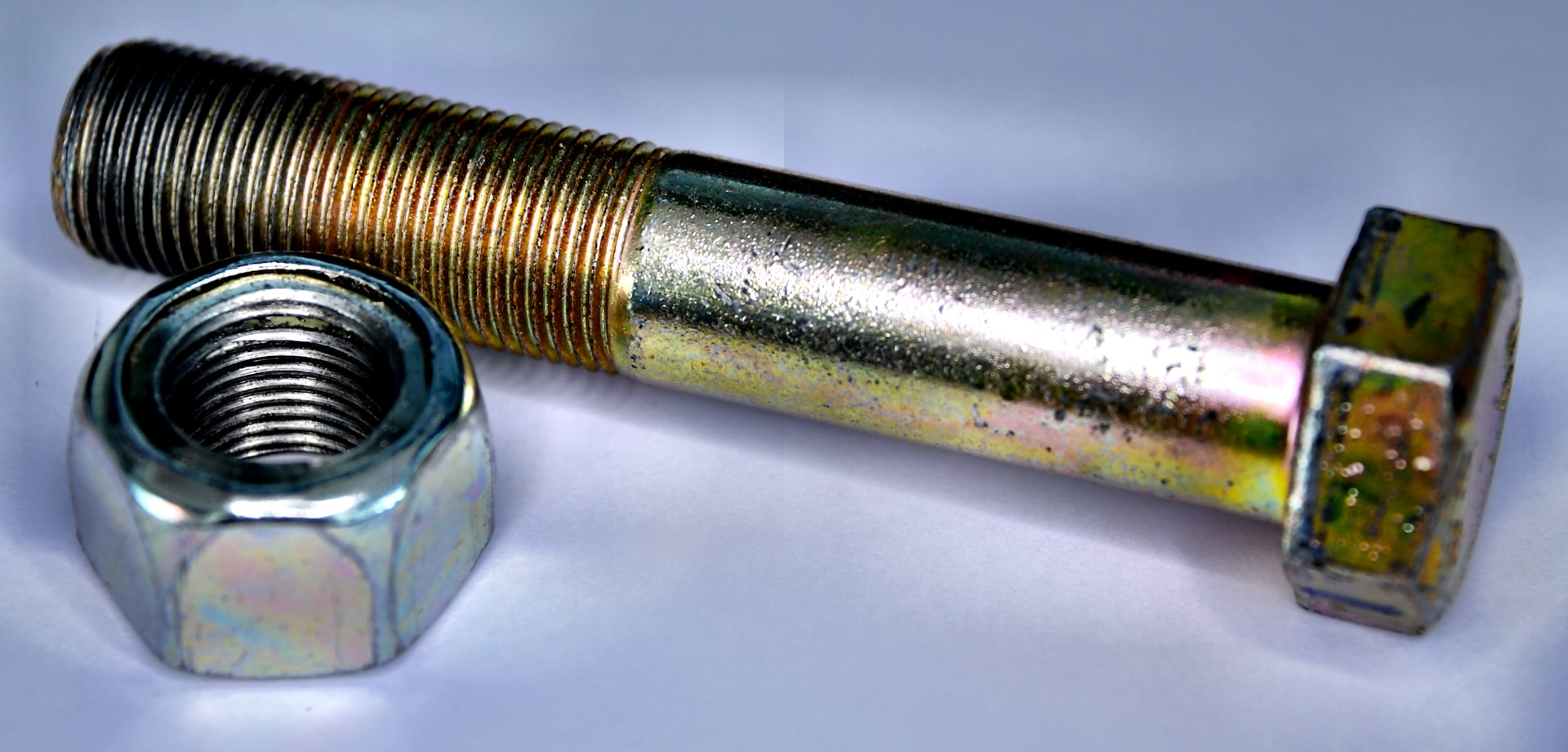 A metallic bolt and nut are placed on a white surface. The bolt has a hexagonal head and is positioned parallel to the nut, which is similarly hexagonal and threaded. Both objects have a shiny metallic surface with slight colorful reflections due to the lighting.