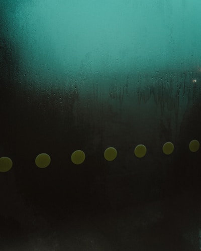 A surface covered in condensation with droplets and streaks forming patterns. A row of evenly spaced yellow-green circles runs horizontally across the lower half of the image, standing out against a gradient background that transitions from dark to teal.