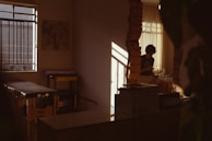 A dimly lit room with a person sitting by a window, partially illuminated by soft, warm light. The room has a massage table, some equipment, and artwork on the wall. Shadows cast interesting patterns, adding depth to the serene space.